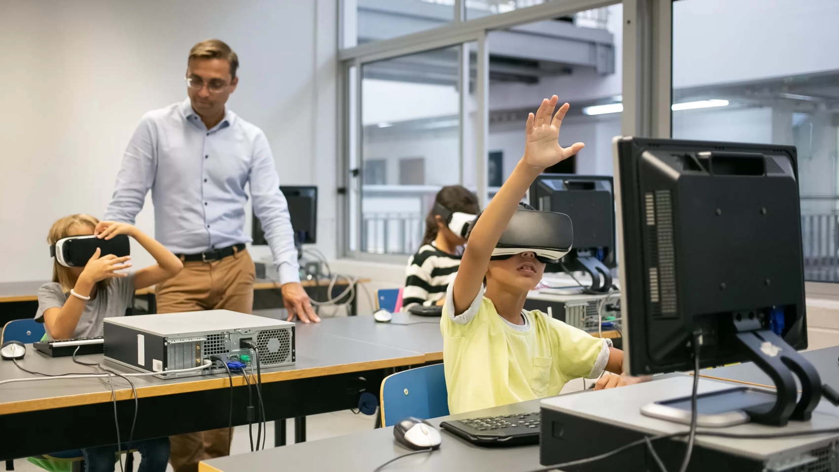 Group of multiethnic kids wearing VR headsets and teacher watching them. Adorable mixed-raced children learning virtual reality and sitting at computer lesson. Informatics and education concept